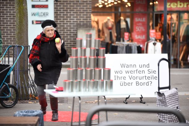 Alte Frau wirf einen gelben Tennisball gegen den Büchsenturm. Daneben steht die Fragentafel: "Wann und wo werfen Sie Ihre Vorurteile über Bord?" 