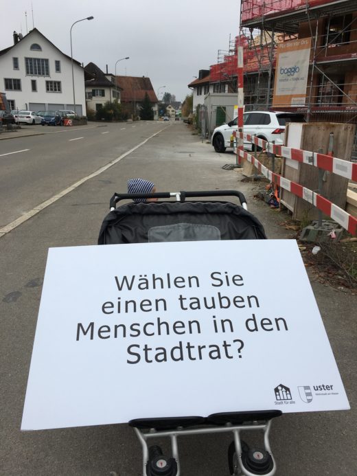 Ein Doppelkinderwagen auf der Strasse. Darauf liegt die Fragentafel: Wählen Sie einen tauben Menschen in der Stadtrat?": "