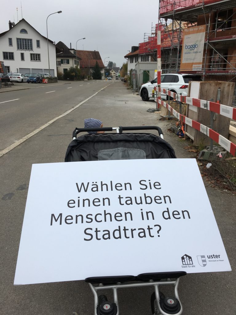 Ein Doppelkinderwagen auf der Strasse. Darauf liegt die Fragentafel: Wählen Sie einen tauben Menschen in der Stadtrat?": "