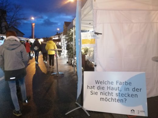 Ustermarkt am Abend. An einem Markstand ist eine Fragentafel angelehnt: "Welche Farbe hat die Haut, in der Sie nicht stecken möchten?"t