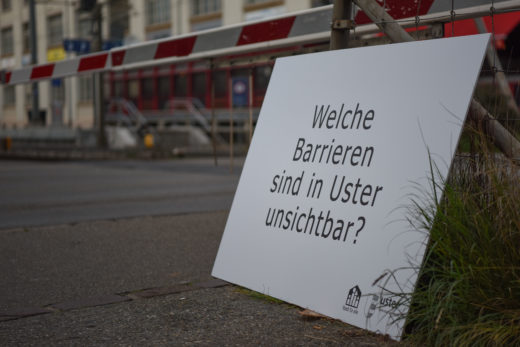 Eine grosse Tafel steht vor der Barriere am Bahnhof Uster. Darauf steht die Frage: "Welche Barrieren sind in Uster unsichtbar?"