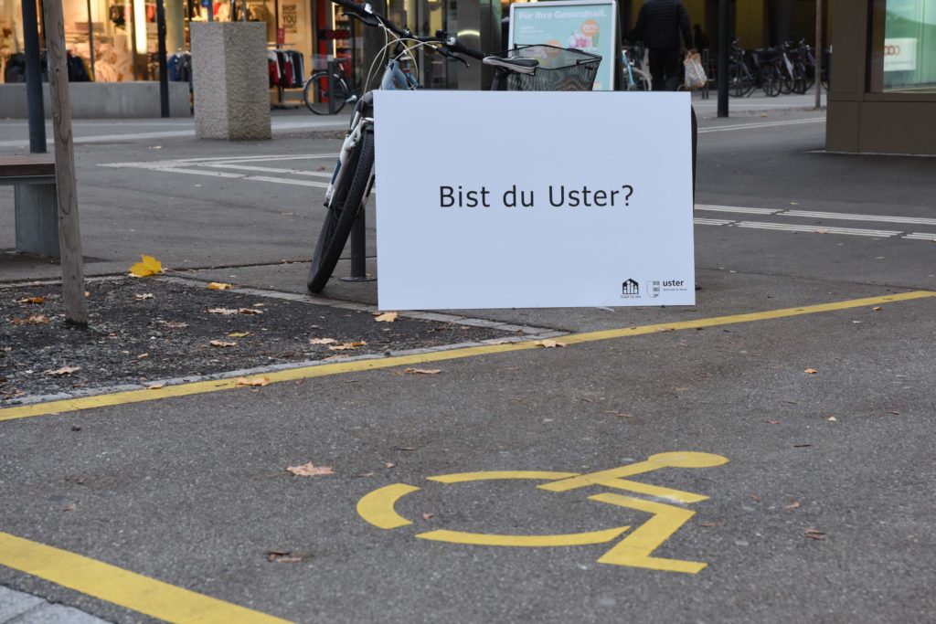 Parkplatz mit einem Reservationszeichen für Menschen mit einer Behinderung. Dahinter steht die Fragentafel: "Bist du Uster?"