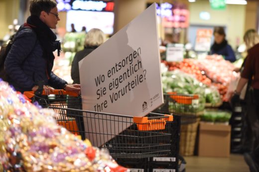 Fragetafel wird von einer Frau im Einkaufswagen durch die Migros geschoben. Auf der Tafel steht: "Wo entsorgen Sie eigentlich Ihre Vorurteile?