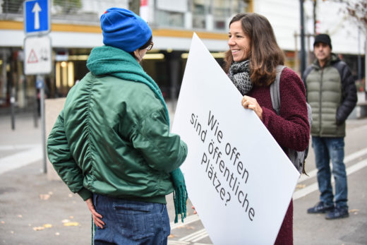 Zwei Frauen im Gespräch. Eine FRau hält die Fragentafel: Wie öffentlich sind öffentliche Plätze?