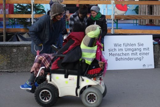 Frau im Rollstuhl im Gespräch mit Lena Estermann von der Zuhörerei. Im Hintergrund die Fragentafel: Wie fühlen Sie sich im Umgang mit Mensche mit eine Behinderung?"