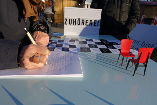 Nahaufnahme einer Hand, die aufschreibt. Auf dem Tisch die Anschrift Zuhörerei und zwei kleine rote Stühle