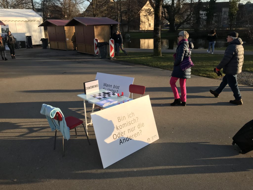 Ein Tisch mit zwei Stühlen im Stadtpark. Fragentafeln an den Tisch gelehnt. Auf der Tafel steht: "Bin ich komisch? Oder nur die andern?"