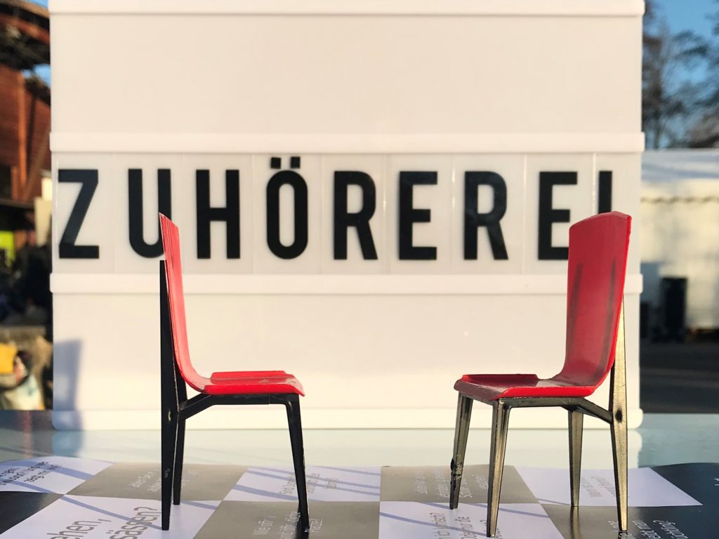 Symbolbild für die Zuhörerei. Schriftzug Zuhörerei, davor zwei kleine rote Puppenstühle.