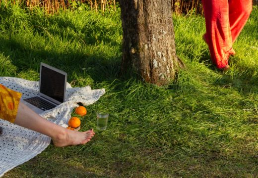 Ein grüner Rasen im Garten. Eine Bein streckt sich auf einer Decke aus, daneben ein Computer und zwei Orangen.