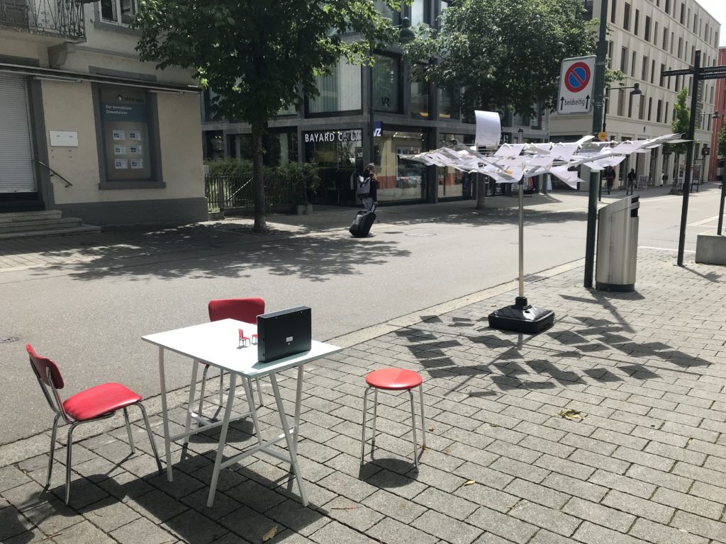 Tisch mit zwei roten Stühlen im Vordergrund, dahinter die Wäschspinne, an der die ausgedruckten Blog-Texte hängen.