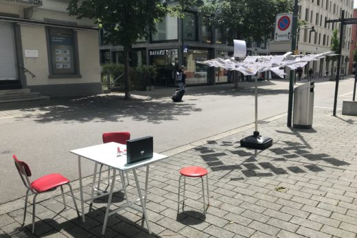 Tisch mit zwei roten Stühlen im Vordergrund, dahinter die Wäschspinne, an der die ausgedruckten Blog-Texte hängen.