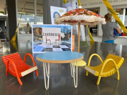 Die Zuhörerei aus Puppenmöbeln: Zwei Sessel, ein Sonnenschirm und ein Tischen sind am See ausgestellt.