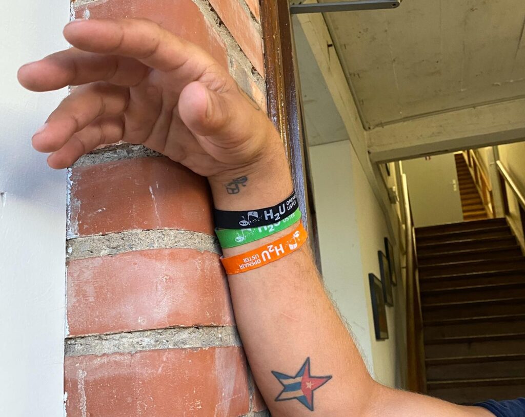 Eine Hand von einem jungen Mann, am Handgelekn drei farbige Bänder mit der Aufschrift H2U Festival.