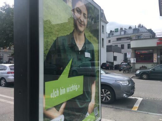 Ein Plakat im öffentlichen Raum bei Bahnhof. Es zeigt eine junge Frau in grünem Pullover. Sie lächelt. Auf dem Plakat ist zu lesen: Ich bin wichtig.