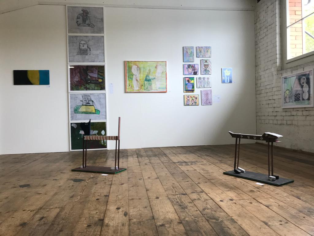 An einer Ausstellungswand hängen verschiedene Kunstwerke. Viele Bilder zeigen eine skizzierte abstrakte Person, die eine Emotion ausdrückt. Ärger, Angst, Langeweile.