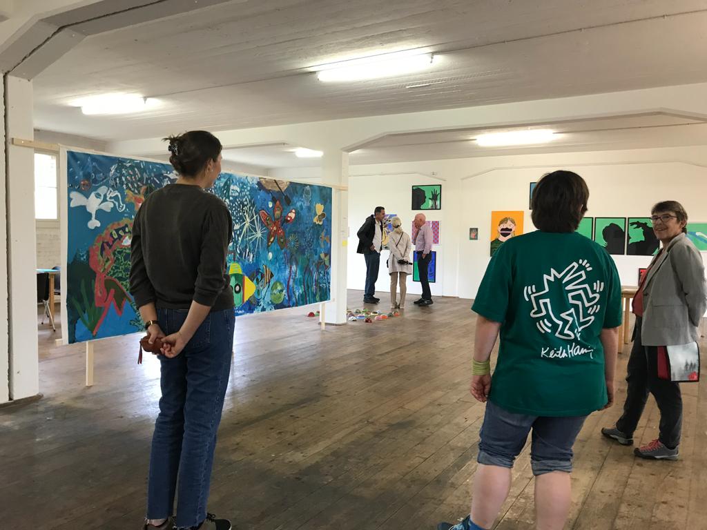 Ein paar Zuschauer*innen schauen sich die Bilderausstellung im K2 auf dem Zeughaus an. Sie stehen vor einem sehr grossen, breiten Bild. Es zeigt ein Meer mit vielen Fischen und Seesternen drin und ist von der Art her eher kindlich gemalt.